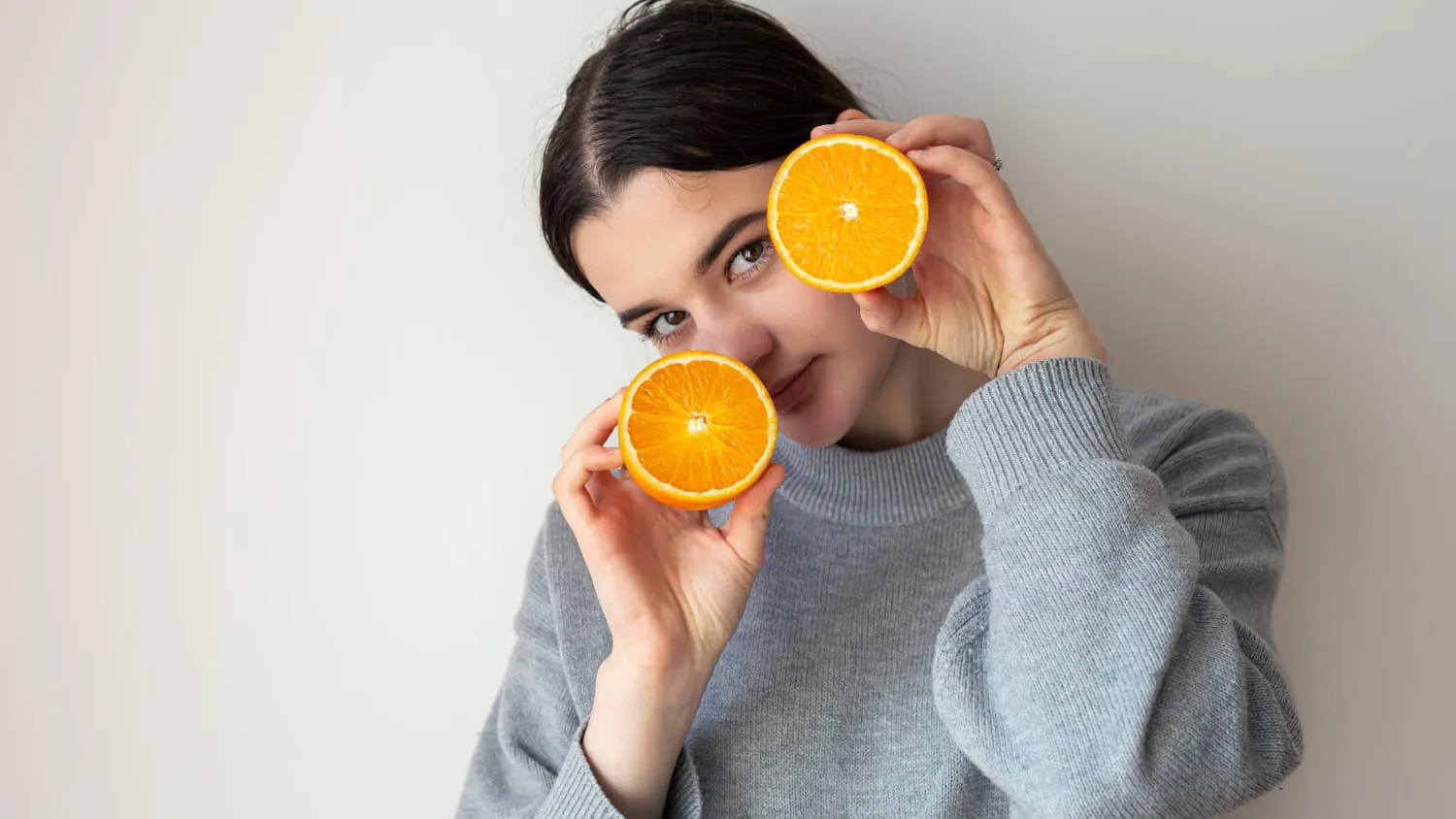 A woman in a knitted sweater holding two orange halves up beside her face