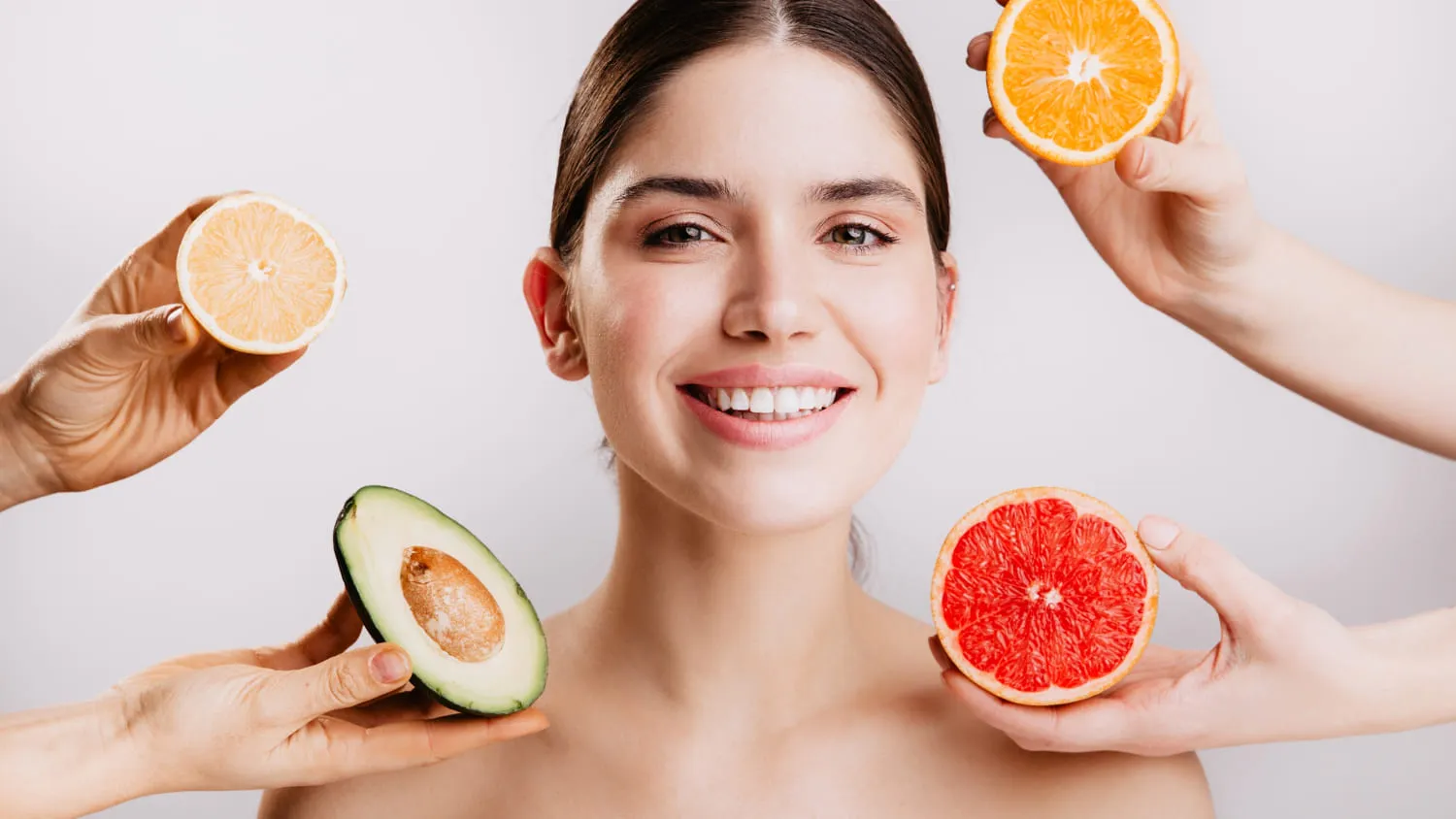 A woman smiling, framed by hands holding halved citrus fruit and an avocado