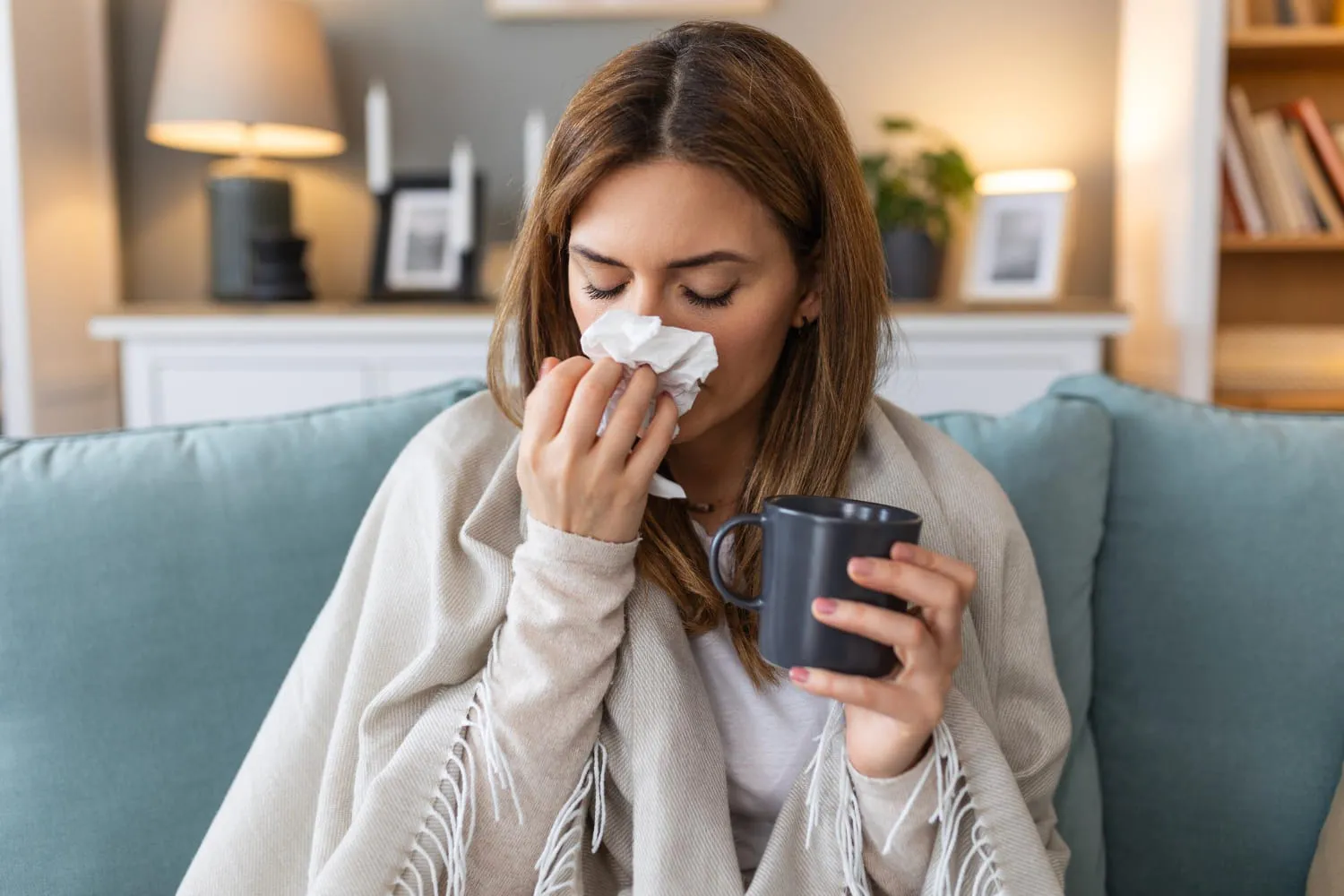 A woman wrapped in a blanket on a sofa at home, holding a mug and a tissue